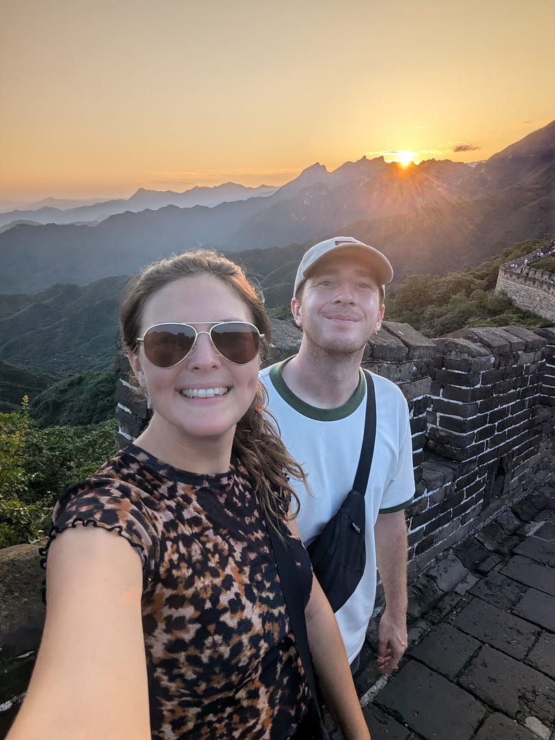 Two Brits on the Go standing on the Great Wall of China with the winding wall and mountain landscape in the background.